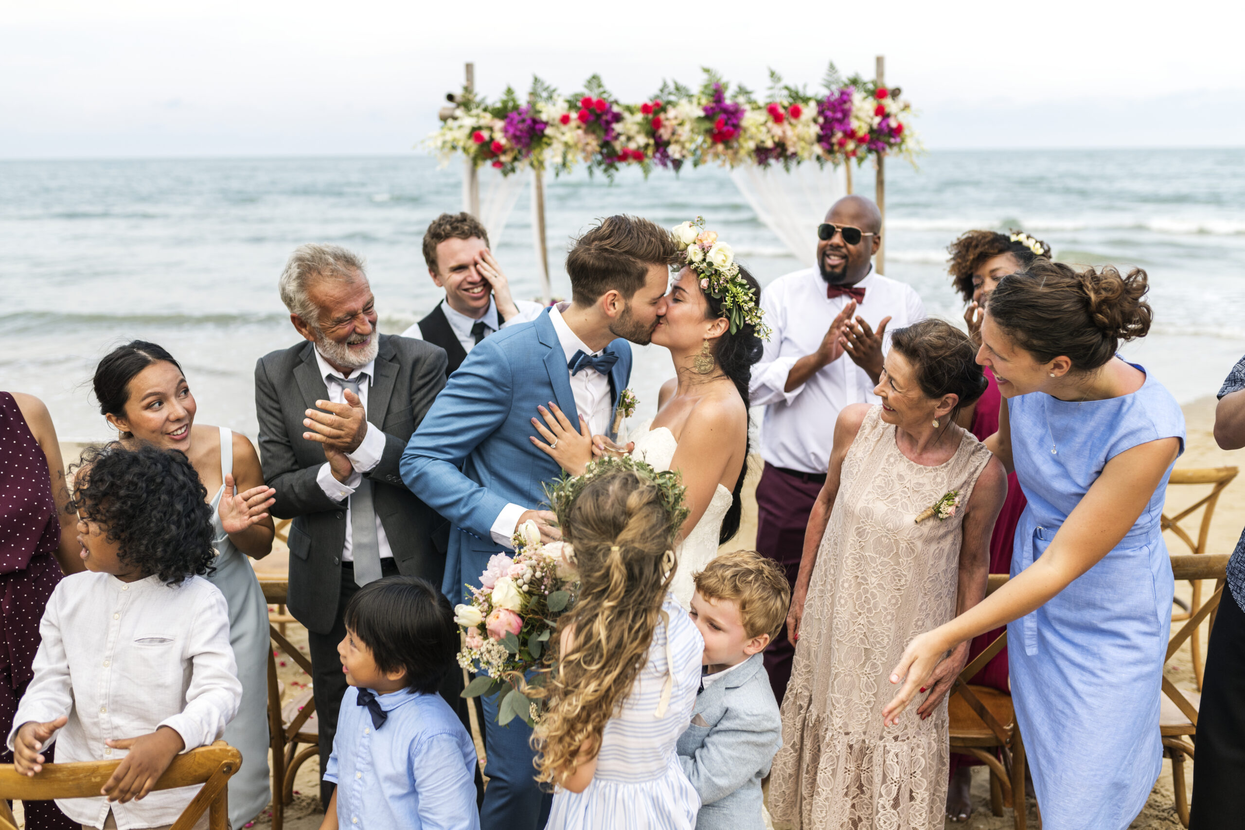 Couple kissing at beach wedding ceremony, surrounded by joyful guests and family.